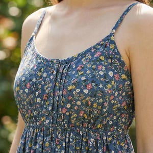 May include: A blue floral print sundress with thin straps. The dress features a button-down front and a gathered skirt. The floral pattern includes small white, yellow, and pink flowers on a blue background. The dress is made of a lightweight fabric.