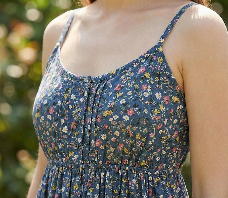 May include: A blue floral print sundress with thin straps. The dress features a button-down front and a gathered skirt. The floral pattern includes small white, yellow, and pink flowers on a blue background. The dress is made of a lightweight fabric.