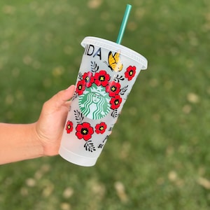 May include: A white reusable cup with a floral design featuring red flowers, green leaves, and a black butterfly. The cup has the word "Vida" written on it and a green straw.