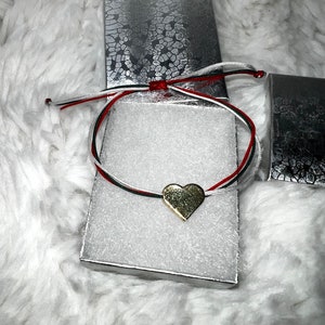 May include: A gold heart charm bracelet with red, black, and white cords. The bracelet is displayed on a white surface inside a silver gift box. The background is a fluffy white material.