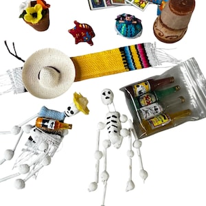 May include: A collection of Day of the Dead themed decorations, including two skeleton figures, a straw hat, a yellow and red woven scarf, a small clay pig, a small doll, a flower pot with flowers, a candle, a pack of Loteria cards, and a bag of miniature bottles of tequila.