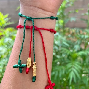 May include: A wrist adorned with two adjustable string bracelets. One is green, the other red. Each bracelet features a cross charm. Additional charms include gold-colored religious figures. The bracelets are tied with knots.