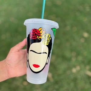 May include: A clear plastic reusable cup with a green straw. The cup has a black and white illustration of Frida Kahlo with red, yellow, and orange flowers in her hair.