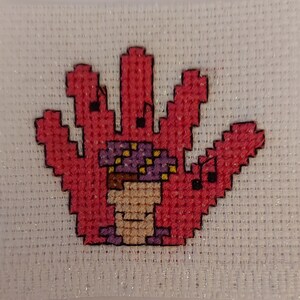 May include: A pink hand-shaped cross-stitch with a purple and yellow mushroom in the centre.