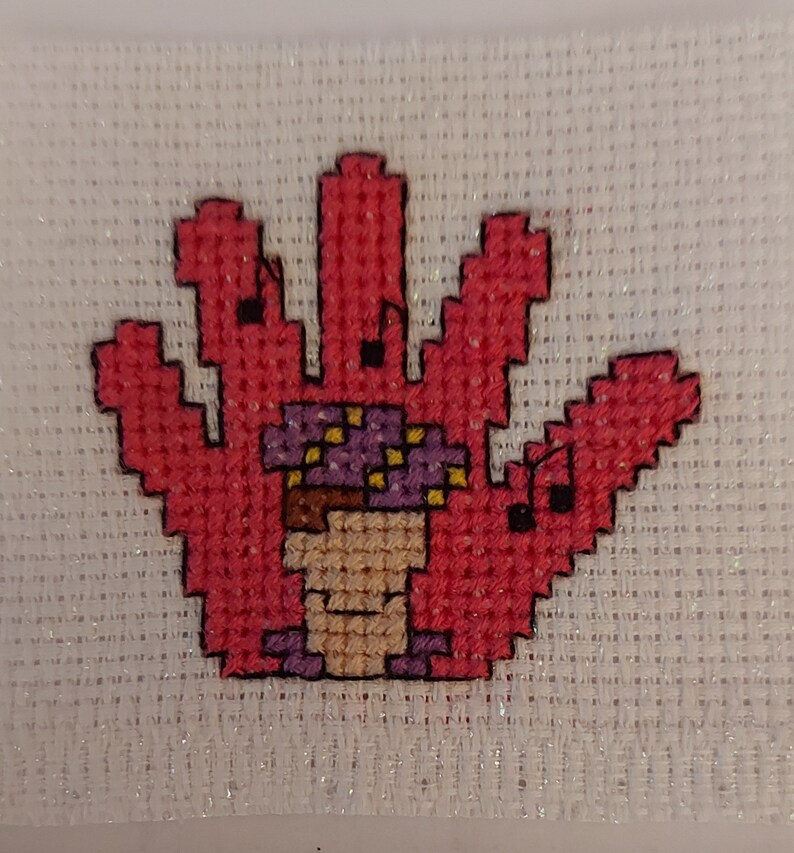 May include: A pink hand-shaped cross-stitch with a purple and yellow mushroom in the centre.