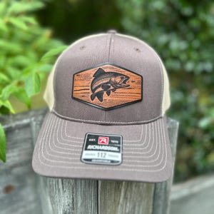 May include: A brown and tan trucker hat with a fish patch. The patch is a brown hexagon with a black fish illustration. The hat has a curved brim and a mesh back. The tag reads "RICHARDSON 112".