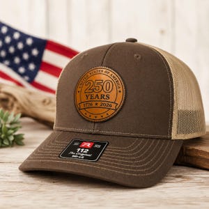 May include: A brown baseball cap with a tan mesh back and a circular leather patch on the front. The patch features the text United States of America, 250 Years, and 1776-2026. A black sticker on the brim displays the text 112 The Original MD-LG.