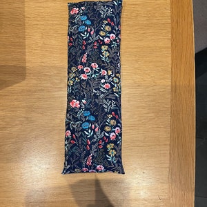 Pode incluir: Uma almofada longa e retangular de tecido azul marinho com um padr&atilde;o floral em tons de rosa, vermelho, azul e amarelo. A almofada &eacute; recheada com arroz e pode ser aquecida no microondas para uma terapia de calor reconfortante.