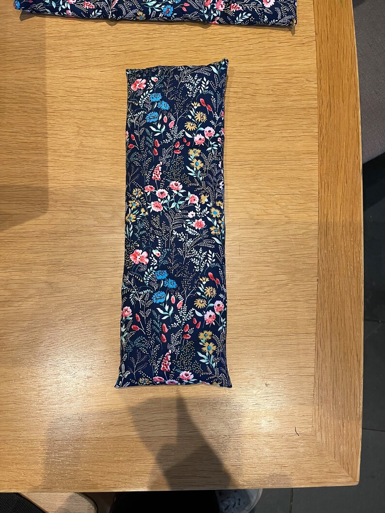 Pode incluir: Uma almofada longa e retangular de tecido azul marinho com um padr&atilde;o floral em tons de rosa, vermelho, azul e amarelo. A almofada &eacute; recheada com arroz e pode ser aquecida no microondas para uma terapia de calor reconfortante.
