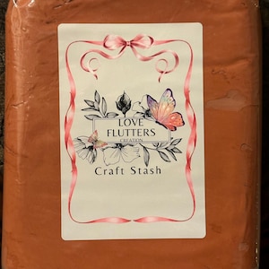 May include: A rectangular block of terracotta-colored clay sealed in clear plastic packaging. A white label features a pink ribbon border, floral illustrations, a butterfly, and the text LOVE FLUTTERS CREATION Craft Stash.