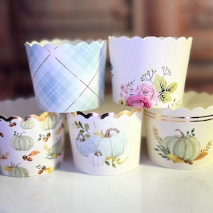 May include: Set of six cupcake liners with scalloped edges. The liners feature different designs, including a blue plaid pattern, a floral pattern with pink roses, and a pumpkin pattern with gold accents.