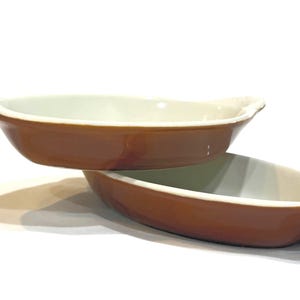 May include: Two brown oval baking dishes with white interiors and small handles. The dishes are stacked, with one slightly above the other. The background is white.