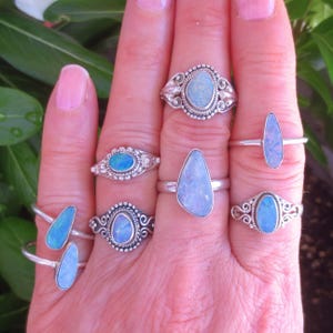 May include: A collection of silver rings with opal stones in various shapes and designs. The rings feature oval, teardrop, and elongated opal stones, showcasing a range of blue and white hues. Some rings have ornate silver detailing.