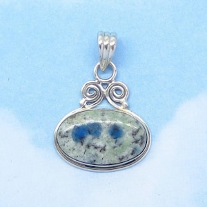 May include: Oval pendant with a silver-colored bail and decorative swirls. The pendant features a light green stone with blue and black speckles, set in a silver-colored bezel. The background is a light blue.