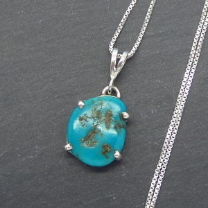 May include: A turquoise pendant necklace with a silver chain. The pendant is a smooth, oval-shaped stone with a dark blue hue and brown markings. The stone is set in a silver bezel with four prongs.