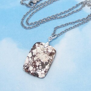 May include: A rectangular pendant featuring a mottled pattern of dark brown and creamy white stone, suspended from a silver-toned metal chain. The pendant has a smooth, polished surface and is attached to the chain by a simple silver-toned metal bail.