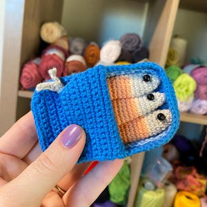 May include: A crocheted blue can with a lid open, revealing three crocheted fish inside. The fish are multi-coloured with white, orange, and grey stripes. Each fish has two black eyes.