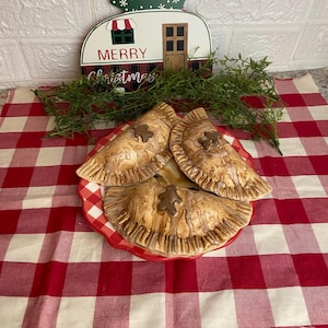 May include: Three homemade gingerbread hand pies on a red and white checkered tablecloth. The pies are topped with a brown glaze and a gingerbread man cutout.