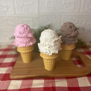 Pu&ograve; includere: Tre palline di gelato testurizzato in coni cialda, che includono una pallina rosa, una bianca e una marrone. I coni sono disposti in fila, ognuno con un colore distinto, mostrando un aspetto realistico di dessert ghiacciati.