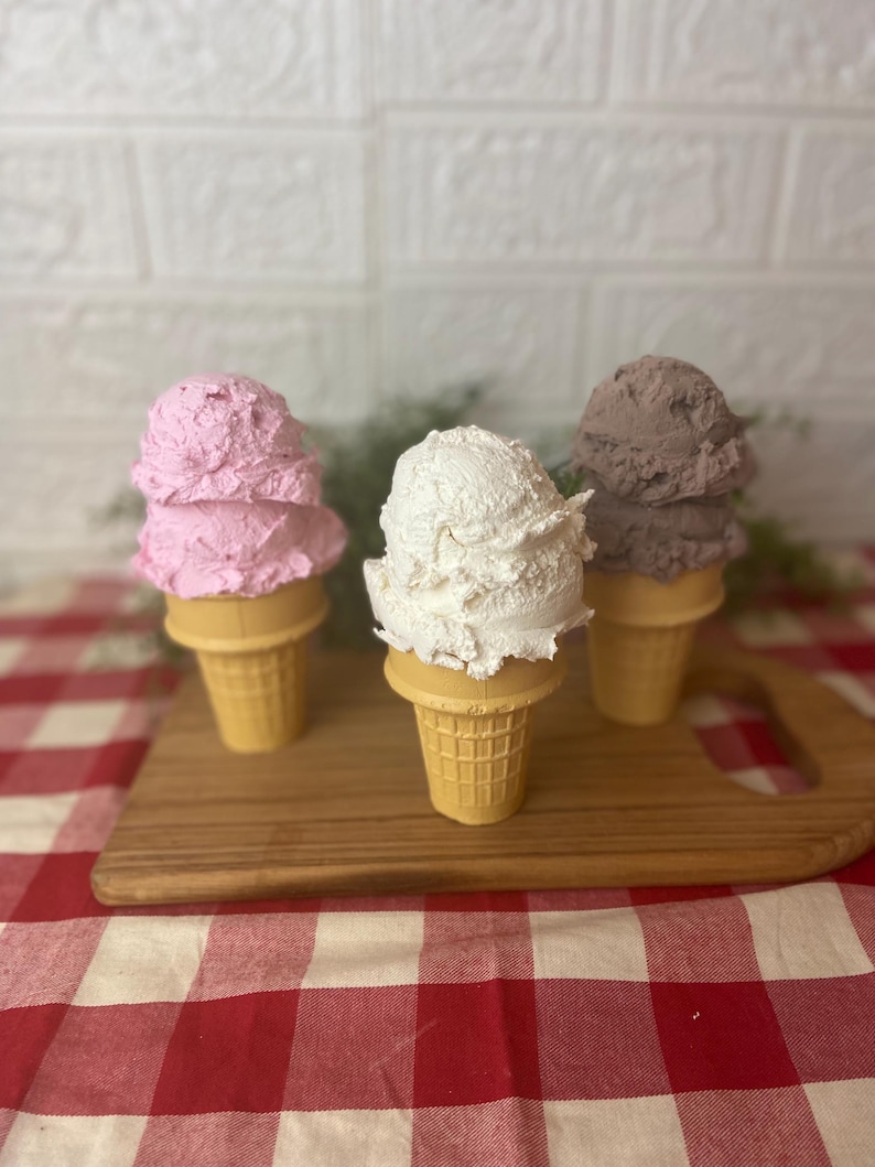 Pu&ograve; includere: Tre palline di gelato testurizzato in coni cialda, che includono una pallina rosa, una bianca e una marrone. I coni sono disposti in fila, ognuno con un colore distinto, mostrando un aspetto realistico di dessert ghiacciati.