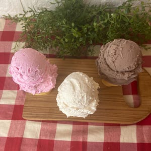 Pu&ograve; includere: Tre palline di gelato nei colori rosa, bianco e marrone, ognuna servita in un cono. Le palline sono disposte su un tagliere di legno, con la pallina rosa a sinistra, la pallina bianca al centro e la pallina marrone sul lato destro.