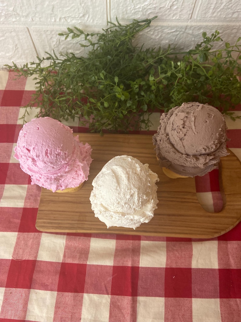 Pu&ograve; includere: Tre palline di gelato nei colori rosa, bianco e marrone, ognuna servita in un cono. Le palline sono disposte su un tagliere di legno, con la pallina rosa a sinistra, la pallina bianca al centro e la pallina marrone sul lato destro.