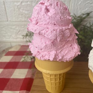 Pu&ograve; includere: Un cono gelato decorativo con due palline sovrapposte di gelato rosa testurizzato su un cono cialda giallo. La superficie delle palline rosa appare densa e montata, mentre il cono presenta un distinto motivo a griglia.