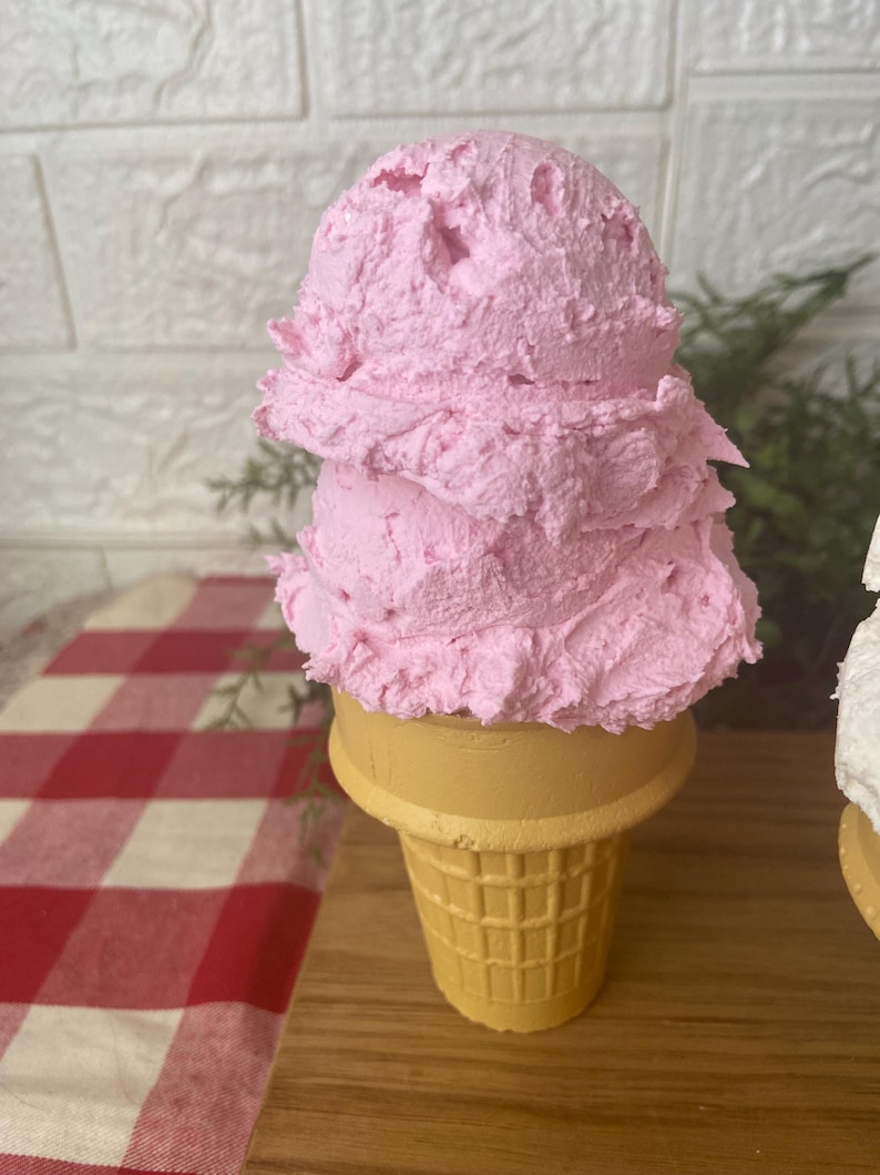 Pu&ograve; includere: Un cono gelato decorativo con due palline sovrapposte di gelato rosa testurizzato su un cono cialda giallo. La superficie delle palline rosa appare densa e montata, mentre il cono presenta un distinto motivo a griglia.