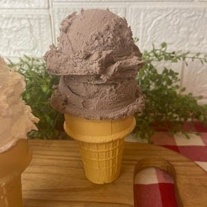Pu&ograve; includere: Una pallina di gelato color marrone cioccolato poggia su un cono cialda giallo chiaro. La superficie strutturata del gelato mostra segni distinti di porzionatore e una forma arrotondata, mentre il cono presenta un classico motivo a griglia.