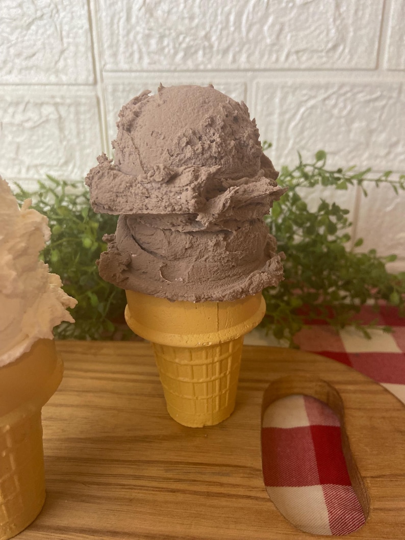 Pu&ograve; includere: Una pallina di gelato color marrone cioccolato poggia su un cono cialda giallo chiaro. La superficie strutturata del gelato mostra segni distinti di porzionatore e una forma arrotondata, mentre il cono presenta un classico motivo a griglia.