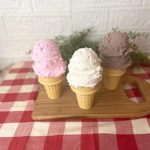 Pu&ograve; includere: Tre palline di gelato testurizzato in coni cialda color marrone chiaro. Una pallina &egrave; rosa chiaro, una &egrave; bianco brillante e una &egrave; di un grigio-marrone tenue. Le palline hanno una consistenza densa e montata, disposte in fila su un tagliere di legno.