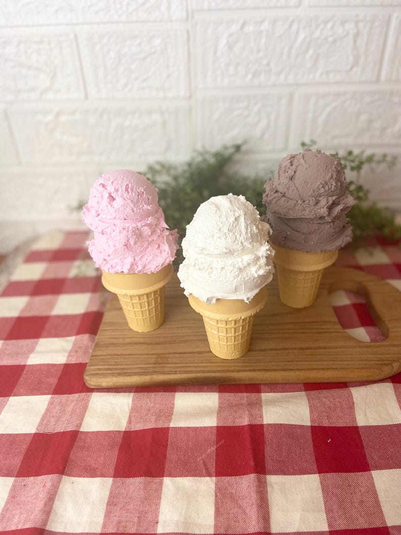 Pu&ograve; includere: Tre palline di gelato testurizzato in coni cialda color marrone chiaro. Una pallina &egrave; rosa chiaro, una &egrave; bianco brillante e una &egrave; di un grigio-marrone tenue. Le palline hanno una consistenza densa e montata, disposte in fila su un tagliere di legno.