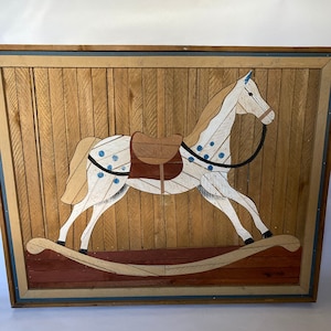 May include: A framed wooden artwork depicting a white rocking horse with blue spots and a brown saddle. The horse is set against a background of wooden planks.