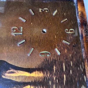 May include: A vintage clock face with a brown background and a wooden frame. The clock face features gold-colored numerals at 12, 3, 6, and 9 o'clock, with gold-colored markers for the other hours. The background depicts a landscape.