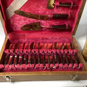 May include: A wooden box with a red velvet interior containing a set of 20 gold-toned flatware with dark wood handles. The box has a brass latch and hinges.