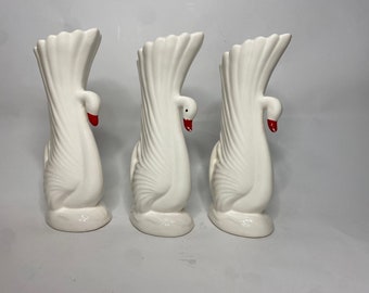 May include: Three white ceramic swan vases with red beaks. The vases are arranged in a row, with the beaks facing forward.