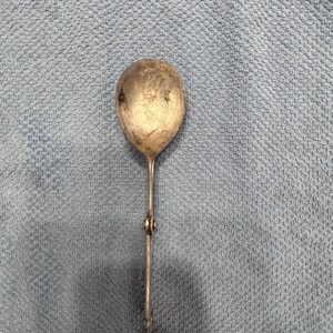 May include: A silver-toned serving spoon with a long, ornate handle. The spoon has a rounded bowl and a decorative design on the handle.