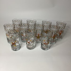 May include: A set of twelve clear drinking glasses. Each glass features a gold rim with a green and red leaf design. The glasses have a central image with a bird and text. The text is in red and the image is in a brown oval.