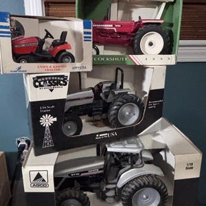 May include: A stack of toy tractors in their original boxes. The top tractor is red with white accents, labeled "Cockshutt 1555." Below is a silver tractor with black tires. The bottom tractor is silver and labeled "White 8710." A red lawn tractor is also visible.