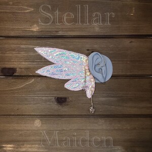 May include: An iridescent, wing-shaped ear cuff featuring light pink fabric with a shimmering, scale-like texture and gold-toned wire detailing. A delicate gold chain hangs from the base, ending in a small, clear, faceted crystal charm.