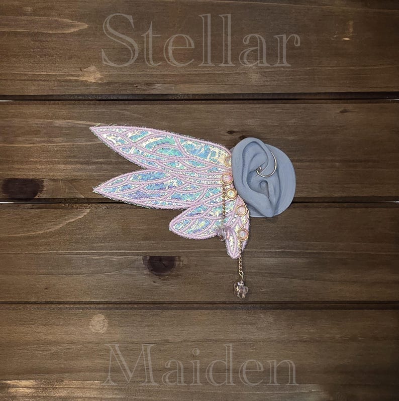 May include: An iridescent, wing-shaped ear cuff featuring light pink fabric with a shimmering, scale-like texture and gold-toned wire detailing. A delicate gold chain hangs from the base, ending in a small, clear, faceted crystal charm.