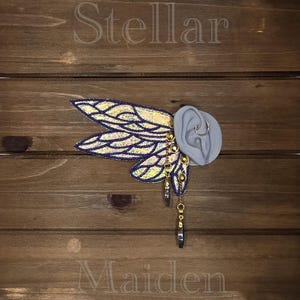 May include: An ear cuff featuring iridescent, wing-shaped fabric attachments with dark purple borders. The piece includes gold-toned metal chains, small circular studs, and two dangling, teardrop-shaped charms attached to a grey ear model.