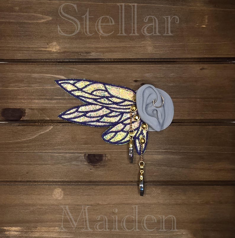 May include: An ear cuff featuring iridescent, wing-shaped fabric attachments with dark purple borders. The piece includes gold-toned metal chains, small circular studs, and two dangling, teardrop-shaped charms attached to a grey ear model.