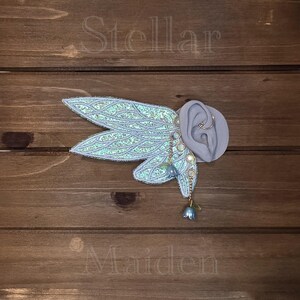 May include: An ear cuff featuring iridescent, light blue, wing-shaped attachments with intricate silver-toned embroidery. The piece includes dangling chains adorned with small, bell-shaped blue charms and circular accents, displayed on a gray ear model.