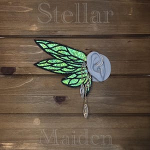 May include: An ear cuff featuring large, iridescent green fairy wings with black embroidered veins. The piece includes a series of small, round, multi-colored cabochons along the edge and two clear, teardrop-shaped crystals dangling from the bottom.