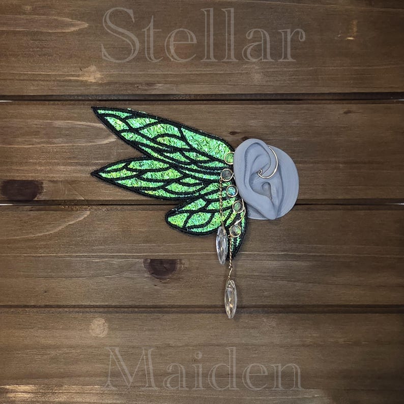May include: An ear cuff featuring large, iridescent green fairy wings with black embroidered veins. The piece includes a series of small, round, multi-colored cabochons along the edge and two clear, teardrop-shaped crystals dangling from the bottom.