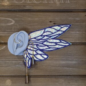 May include: An ear cuff featuring iridescent, wing-shaped embroidery with dark blue stitching. The design includes small, round, light-reflecting beads and two dangling, dark-toned, cone-shaped charms suspended by thin gold-colored metal chains.