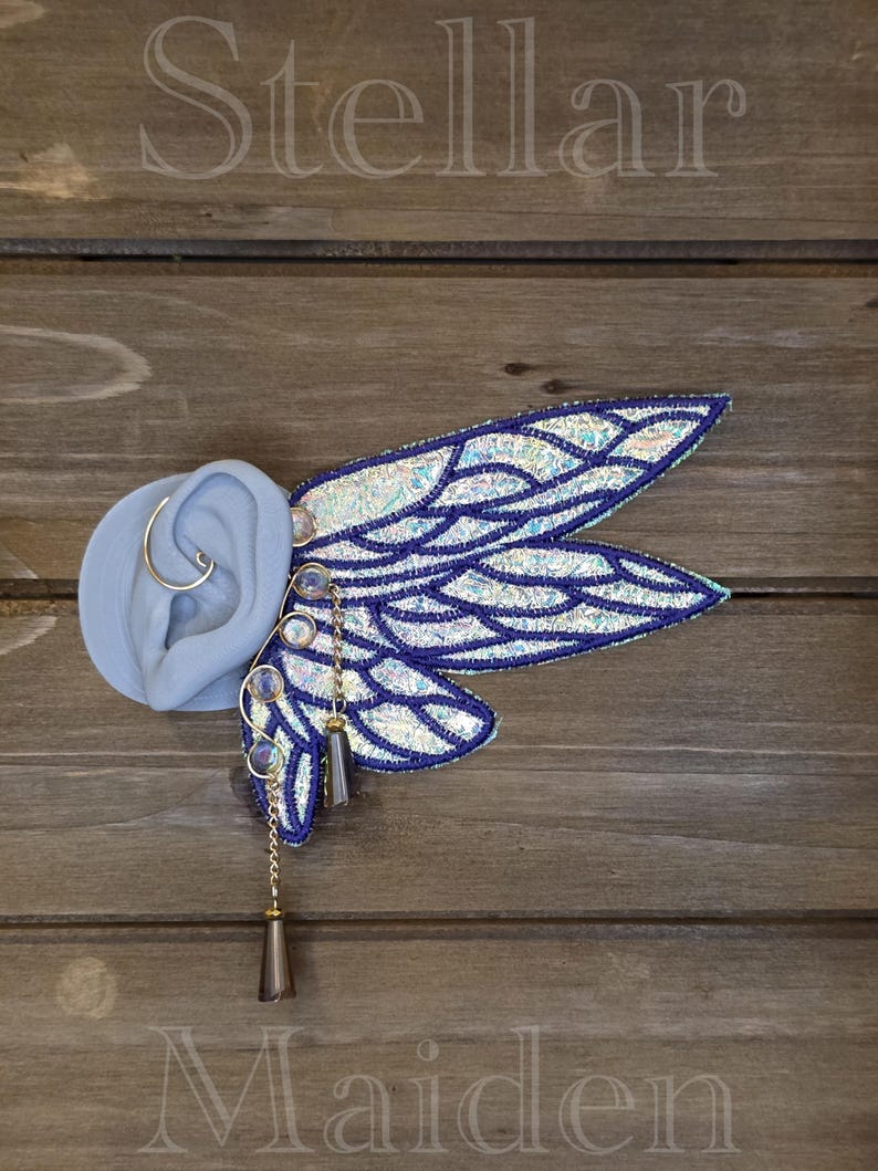 May include: An ear cuff featuring iridescent, wing-shaped embroidery with dark blue stitching. The design includes small, round, light-reflecting beads and two dangling, dark-toned, cone-shaped charms suspended by thin gold-colored metal chains.