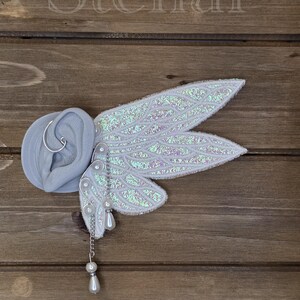 May include: An ear cuff featuring iridescent white fairy wings with a textured surface, attached to a gray ear model. The accessory includes silver-toned wire, small white faux pearls, and a dangling chain with a teardrop-shaped pearl charm.