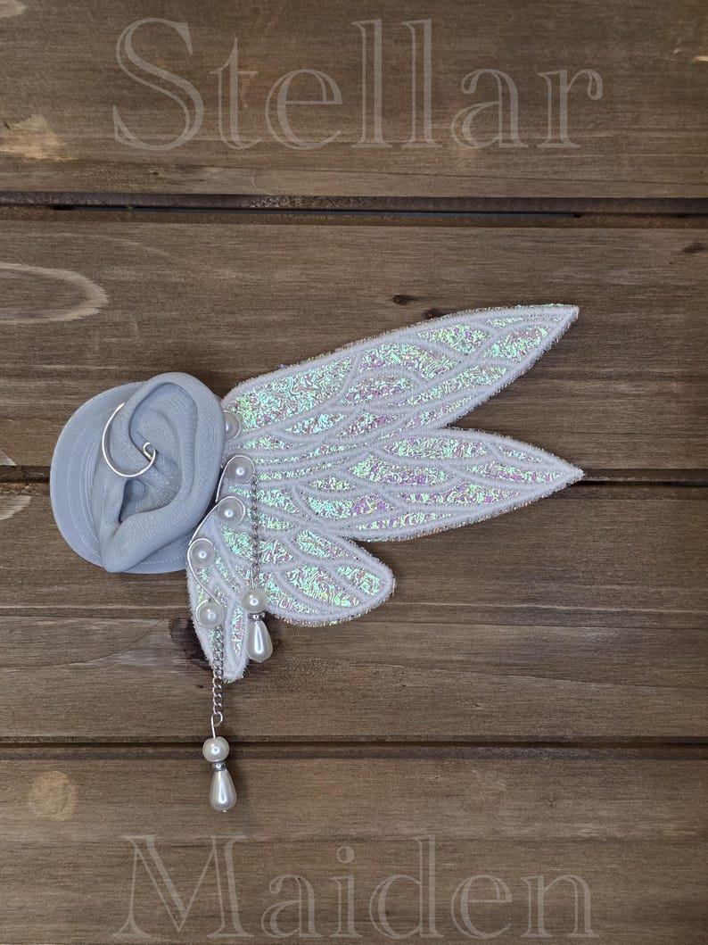 May include: An ear cuff featuring iridescent white fairy wings with a textured surface, attached to a gray ear model. The accessory includes silver-toned wire, small white faux pearls, and a dangling chain with a teardrop-shaped pearl charm.